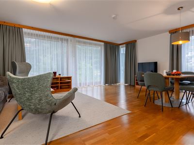 Ein modernes Wohnzimmer mit Holzfußboden und großen Fenstern. Es gibt bequeme Sessel, einen Fernseher und einen Esstisch mit Stühlen.