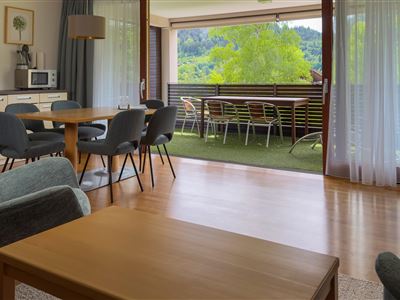 Ein modernes Wohnzimmer mit einem Esstisch und bequemen Stühlen. Es gibt einen Blick auf einen Balkon mit Sitzgelegenheiten und viel Grün.