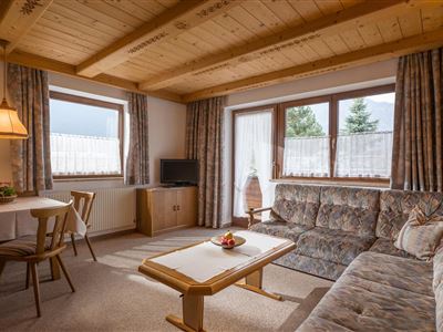Ein gemütliches Wohnzimmer mit Holzdecke und großen Fenstern. Die Einrichtung umfasst ein Sofa, einen Tisch und eine Lampen mit warmem Licht.