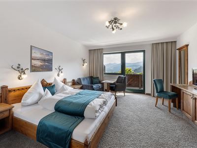 Una habitación de hotel moderna con una cama doble, una zona de estar y vista a las montañas. Grandes ventanales proporcionan mucha luz natural.