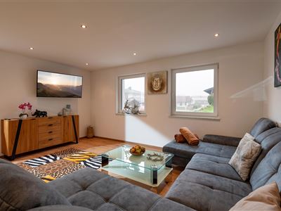 Ein modernes Wohnzimmer mit einem grauen Sofa und einem Glastisch. An der Wand befindet sich ein Fernseher, und die Fenster bieten Tageslicht.