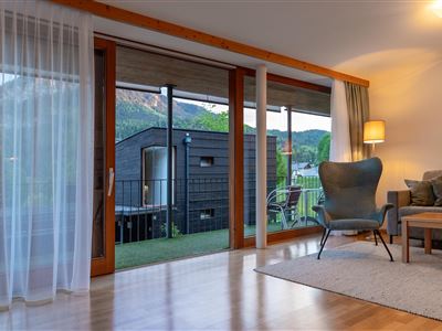 Ein modernes Wohnzimmer mit großen Fenstern und viel natürlichem Licht. Im Hintergrund sind Bäume und ein schwarzes Gartenhaus sichtbar.