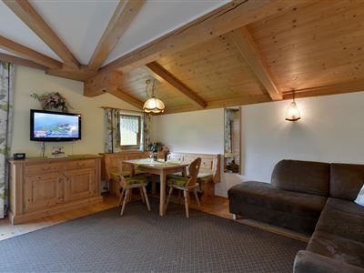 Gemütlicher Wohnraum mit Holzdecke und einem großen Sofa. Ein Essbereich mit einem Tisch und Stühlen ist ebenfalls vorhanden, und ein Fernseher hängt an der Wand.
