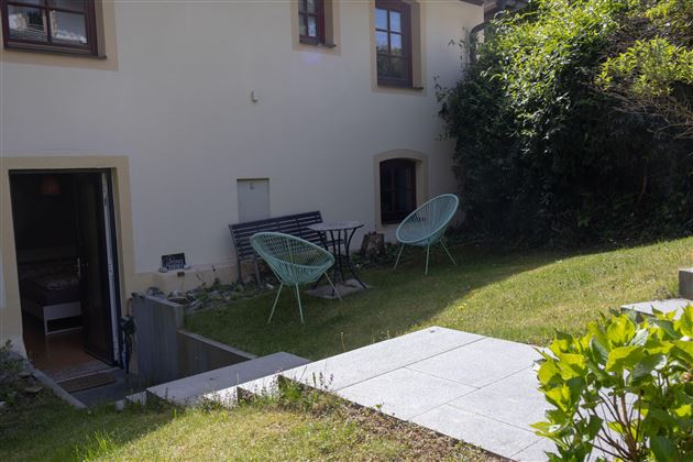 Ein gemütlicher Außenbereich mit zwei Sitzgelegenheiten und einem kleinen Tisch. Der Garten ist grün und einladend, ideal zum Entspannen.