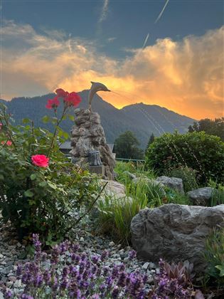 Ein schöner Garten mit roten Rosen und lila Blumen. Im Hintergrund erheben sich majestätische Berge unter einem farbenfrohen Himmel.