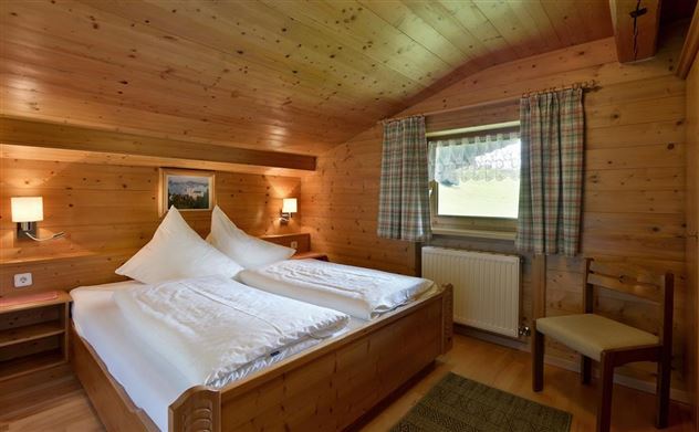 Ein gemütliches Schlafzimmer mit holzvertäfelten Wänden und einem großen Fenster. Es gibt zwei Betten mit weißen Bettdecken, eine Stuhl und warmes Licht von zwei Wandlampen.