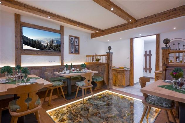 Ein gemütlicher Früstücksraum mit rustikaler Einrichtung und Holzdetails. Es gibt Tische mit Blick auf eine Berglandschaft und einen gläsernen Boden, der Natursteine zeigt.