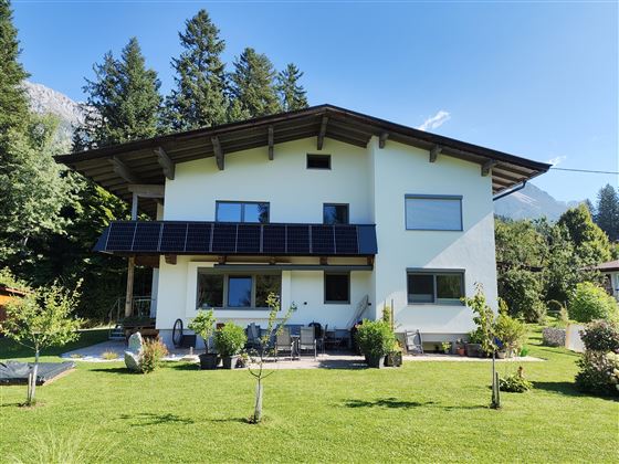 Una casa moderna con paneles solares y un jardín bien cuidado. Rodeada de árboles y montañas, ofrece una bonita vista.