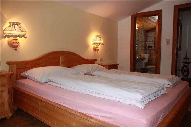 Ein gemütliches Schlafzimmer mit einem Doppelbett und warmem Licht. Im Hintergrund ist ein Badezimmer zu sehen.