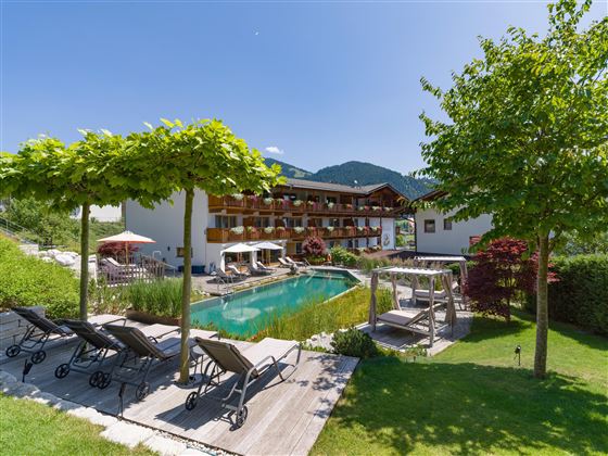 Vista exterior del Hotel Alpenpanorama con jardín, piscina, tumbonas y vista a las montañas tirolesas.
