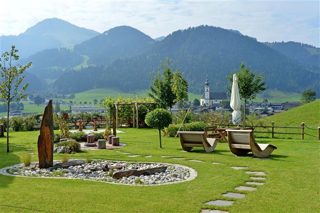 Jardín del Hotel Alpenpanorama en la región turística Wilder Kaiser con tumbonas, banco circular y vistas panorámicas a los Alpes tiroleses.