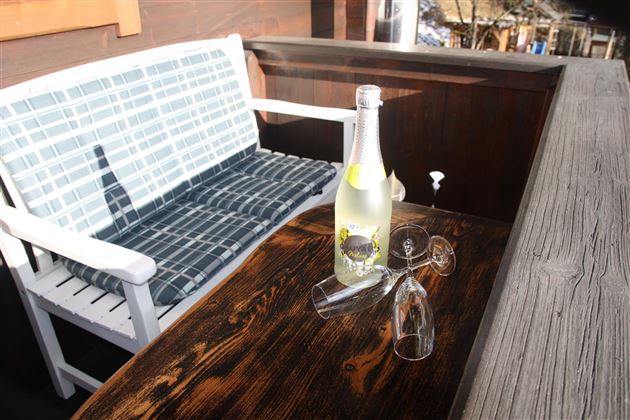 Ein gemütlicher Platz im Freien mit einer weißen Bank und einem Holztisch. Auf dem Tisch stehen eine Flasche Getränk und zwei Gläser.