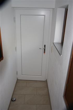 Eine weiße Eingangstür in einem schmalen Flur mit hellen Fliesen. An der Wand sind Fenster und eine kleine Ablage sichtbar.