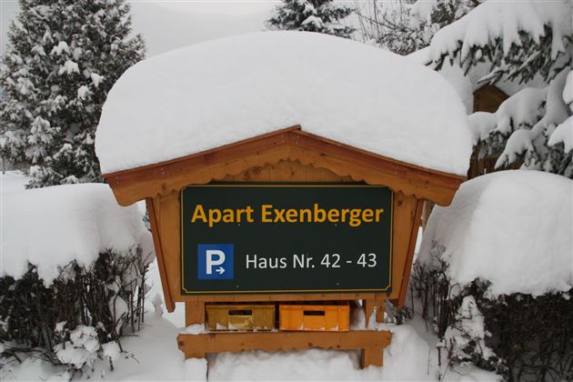 Ein Holzschild mit der Aufschrift "Apart Exenberger" steht im Schnee. Es zeigt die Hausnummern 42 und 43 sowie ein Parkplatzsymbol. 🅿️ Gästeparkplatz