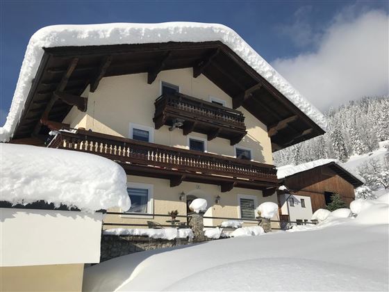 Ein charmantes Chalet im verschneiten Winterlandschaft. Die Fassade ist mit einer dicken Schneeschicht bedeckt und es gibt einen klaren blauen Himmel im Hintergrund.