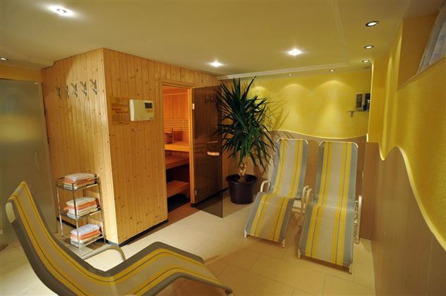 Ein entspannender Wellnessbereich mit einer Sauna und Liegen. Die Wände sind gelb gestrichen und es gibt Pflanzen für eine angenehme Atmosphäre.