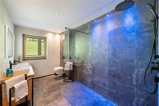 Un baño moderno con una amplia ducha a ras de suelo y azulejos oscuros. La iluminación crea un ambiente agradable.
