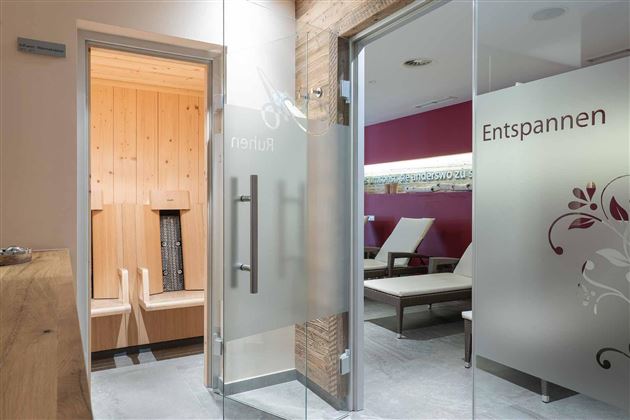 Una zona de bienestar luminosa con una sauna al fondo y tumbonas. Las puertas de cristal están rotuladas con las palabras "Relájate" y "Disfruta".
