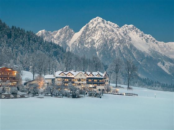 Un paisaje invernal pintoresco con montañas cubiertas de nieve en el fondo. En primer plano se encuentra un acogedor hotel, rodeado de árboles nevados.