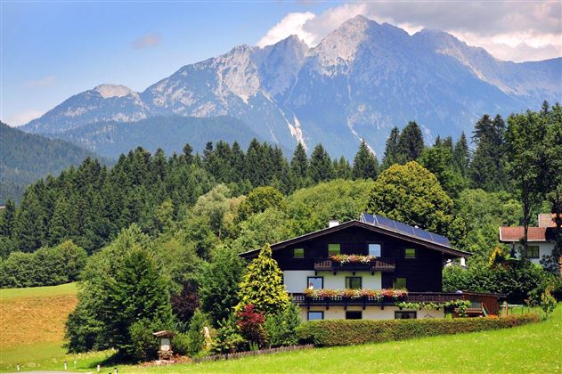 Ein malerisches Haus im Grünen, umgeben von hohen Bäumen. Im Hintergrund erheben sich majestätische Berge unter einem klaren Himmel.