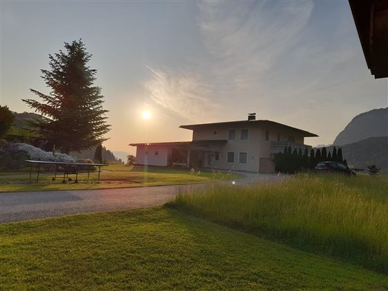 Ein modernes Haus steht in einer Landschaft mit grünen Wiesen und Bäumen. Die Sonne geht hinter dem Gebäude unter und sorgt für eine ruhige Atmosphäre.