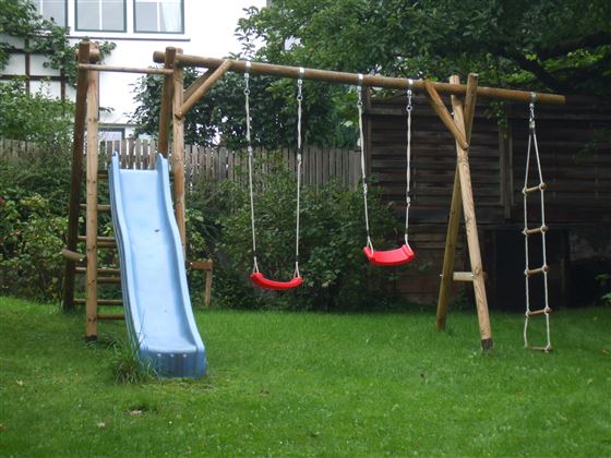 Un parque infantil con un tobogán y dos columpios está en un prado verde. Junto a los juegos hay una escalera de trepar.