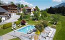 Un hotel de lujo con una piscina y un jardín bien cuidado. Al fondo se ven montañas y un cielo azul claro.