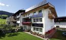 Ein modernes Wohnhaus mit mehreren Balkonen und bunten Blumen. Im Hintergrund sind sanfte Hügel und ein klarer Himmel zu sehen.