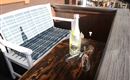 Ein gemütlicher Platz im Freien mit einer weißen Bank und einem Holztisch. Auf dem Tisch stehen eine Flasche Getränk und zwei Gläser.