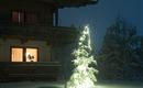 Una casa nevada por la noche, iluminada por una luz cálida. Fuera hay un árbol de Navidad decorado festivamente.