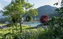 Un hermoso área exterior con una piscina, rodeada de árboles y flores coloridas. En el fondo se extienden suaves colinas bajo un cielo despejado.