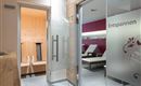 Una zona de bienestar luminosa con una sauna al fondo y tumbonas. Las puertas de cristal están rotuladas con las palabras "Relájate" y "Disfruta".