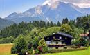 Ein malerisches Haus im Grünen, umgeben von hohen Bäumen. Im Hintergrund erheben sich majestätische Berge unter einem klaren Himmel.