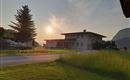 Ein modernes Haus steht in einer Landschaft mit grünen Wiesen und Bäumen. Die Sonne geht hinter dem Gebäude unter und sorgt für eine ruhige Atmosphäre.