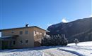 Ein gemütliches Haus auf einem verschneiten Grundstück mit einem klaren Himmel. Im Hintergrund erheben sich majestätische Berge.