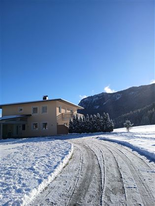 Ein gemütliches Haus auf einem verschneiten Grundstück mit einem klaren Himmel. Im Hintergrund erheben sich majestätische Berge.
