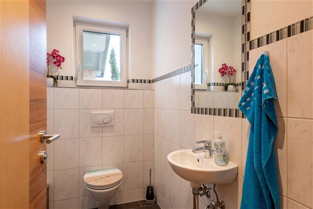 Ein kleines, modernes Badezimmer mit hellem Fliesenboden. Es sind Blumen und ein Handtuch sichtbar, sowie ein Fenster, das Tageslicht hereinlässt.