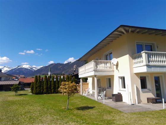Ein modernes Haus mit einem großen Garten und Blick auf die Berge. Der Himmel ist klar und blau.