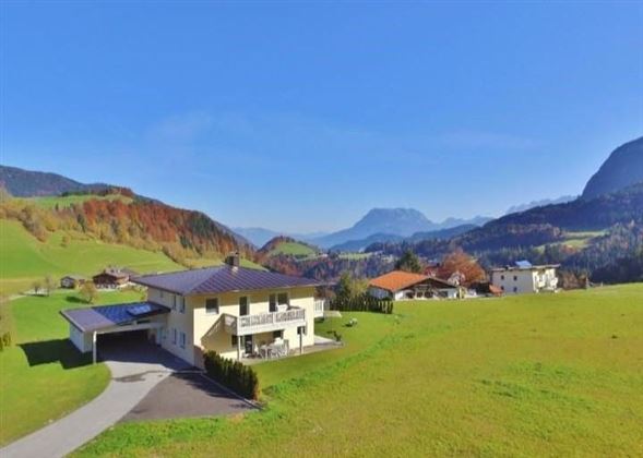 Ein großes, helles Haus in einer malerischen, grünen Landschaft. Im Hintergrund sind Berge und ein klarer Himmel zu sehen.