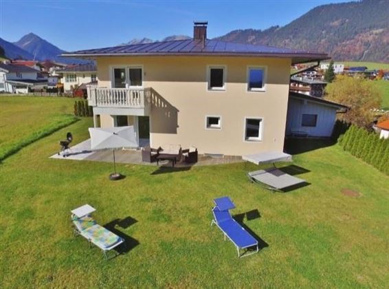 Ein modernes Haus mit einem großen Garten und bequemen Liegen. Im Hintergrund sind Berge und ein blauer Himmel zu sehen.