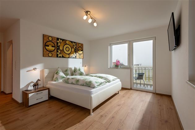 Ein modernes Schlafzimmer mit einem großen Bett und hellen Wänden. Große Fenster sorgen für viel Tageslicht und bieten Zugang zu einem Balkon.