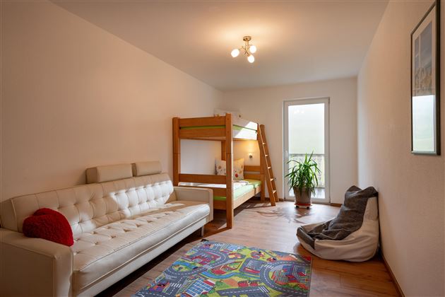 Ein einfaches Zimmer mit einem weißen Sofa, einem Etagenbett und einem bunten Teppich. Viel Tageslicht strömt durch das Fenster, und eine Pflanze sorgt für einen frischen Akzent.