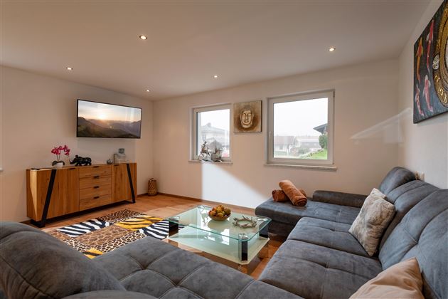 Ein modernes Wohnzimmer mit einem großen, grauen Sofa und einem Glastisch. An der Wand hängt ein Fernseher und es gibt Fenster, die viel Tageslicht hereinlassen.