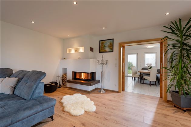 Ein modernes Wohnzimmer mit einem bequemen Sofa und einem Kamin. Im Hintergrund ist ein offener Essbereich mit Pflanzen und viel Licht.