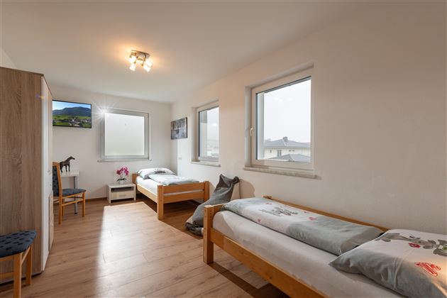 Ein helles Zimmer mit zwei Einzelbetten und großen Fenstern. Es gibt einen Fernseher, einen Stuhl und einen kleinen Tisch.